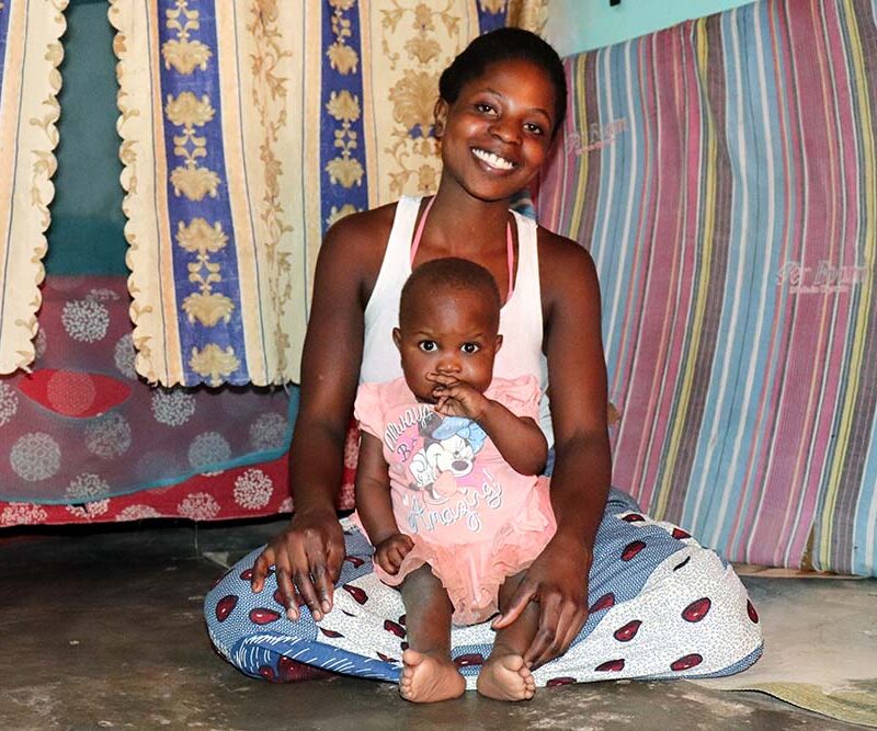 A woman sits smiling on the floor with her baby on her lap.