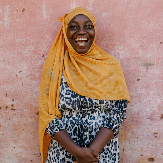 A young woman wearing a yellow headscarf and leopard print dress laughs in front of a pink wall